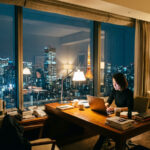 夜景が見えるホテルのデスクでノートPCを開き、集中して仕事をするノマドワーカー