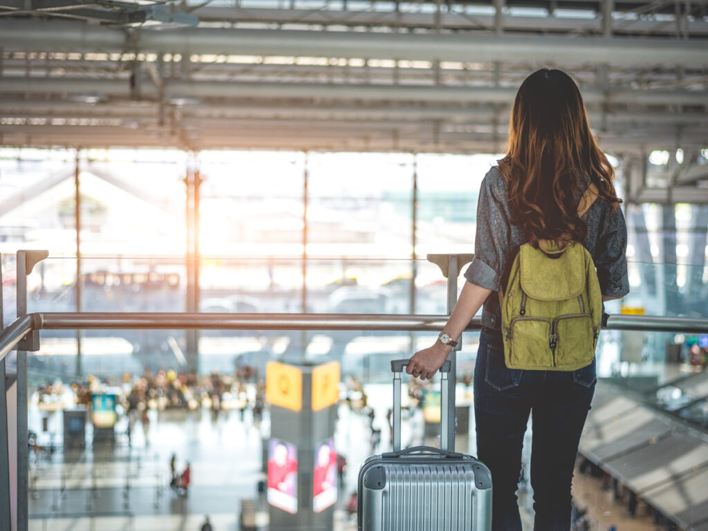 広い空港ターミナルでスーツケースを引きながら出発ロビーを見下ろすバックパック姿の女性