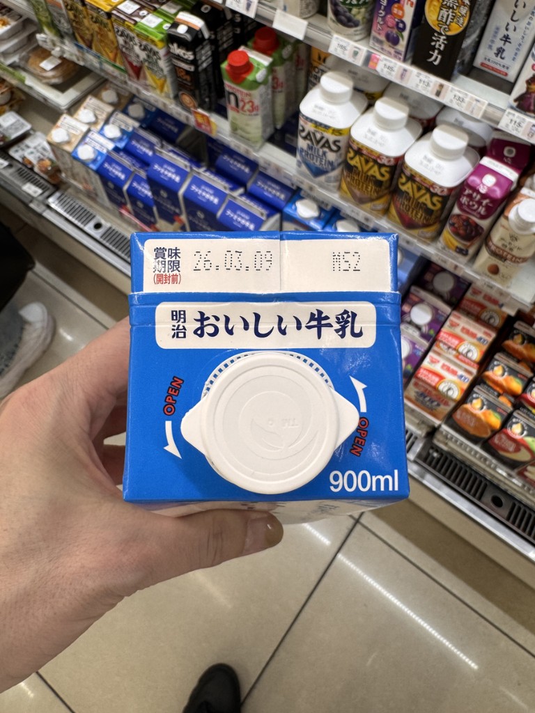 900ml Meiji milk carton held in front of a Japanese convenience store drink chiller with SAVAS protein drinks and other beverages