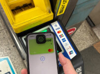 iPhone showing Wise card in Apple Wallet held to a Japan train ticket gate contactless reader with Tap to ride signage.